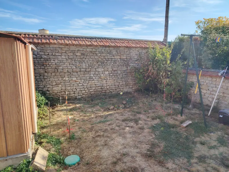 Angle du jardin en herbe sèche devant un mur en pierre coiffé de tuiles, avant la construction