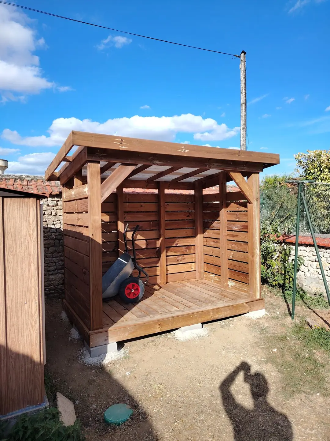 Abri à bois sur mesure en bois, bardage à claire-voie et plancher, posé sur plots béton devant un mur en pierre