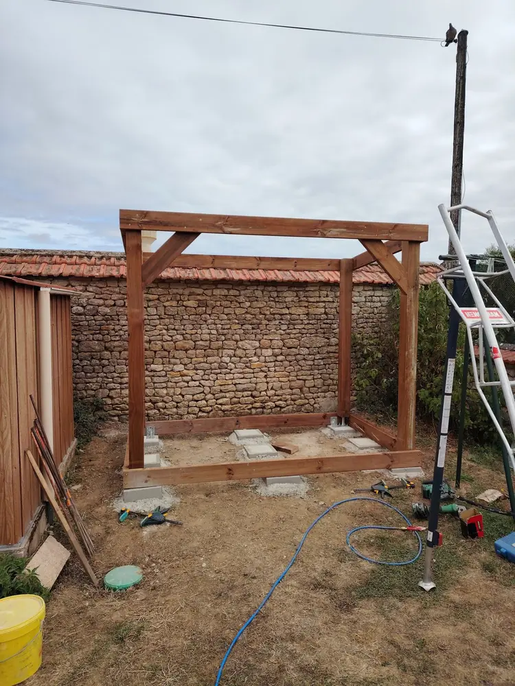 Ossature bois de l’abri en cours de montage, posée sur des plots béton devant le mur en pierre