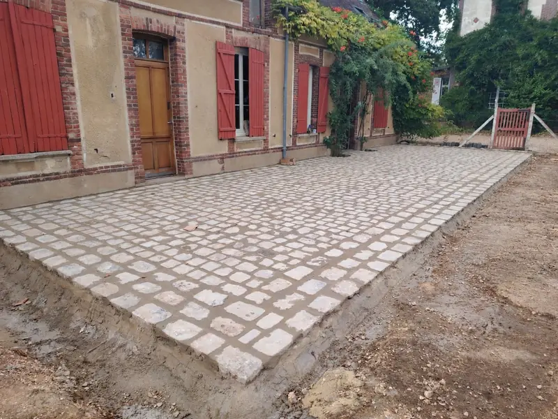 Terrasse en pavés anciens terminée devant une maison aux volets rouges, ceinture de gros pavés au premier plan