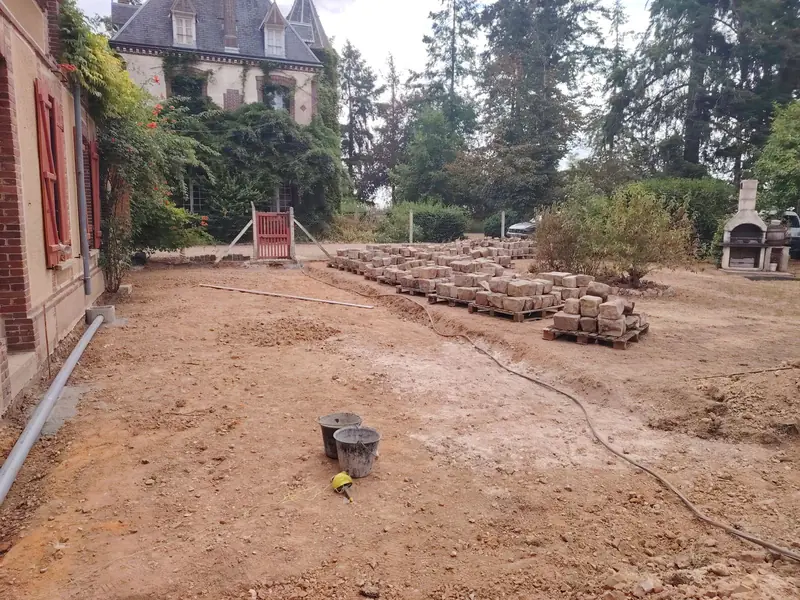 Terrain décaissé le long de la façade, pavés anciens rangés sur palettes au fond