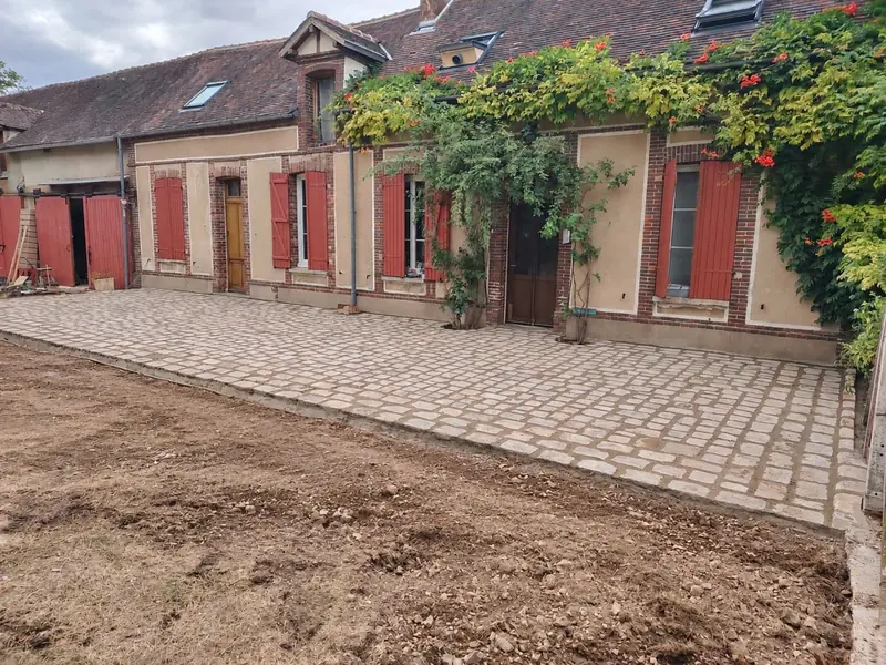 Vue d’ensemble de la terrasse en pavés anciens terminée devant la maison, gazon à refaire au premier plan