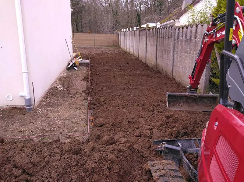 Mini-pelle VB Jardin nivelant de la terre végétale le long d’une clôture